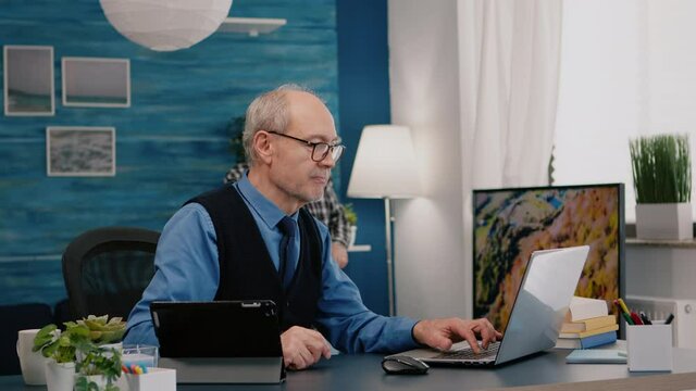 Senior man using laptop and tablet in same time working in office living room at project. Busy focused old employee writing, searching, analysing reports while elderly wife watching tv in background