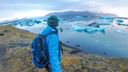A young man wearing a blue jacket and a hiking backpack gazes on the glacier lagoon from a grass shore. He is taking a selfie with a stick. Drifting ice bergs in the lagoon. Peaceful and quiet.  © Chris
