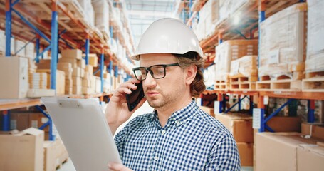 Warehouse manager in glasses and white helmet standing on background of rows of parcels and talking on mobile pnone holding paper fodler.