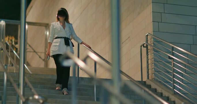 Stylish young woman walking down the stairs. Model wearing trendy white and black outfit, long blazer with many pockets, fashionable black belt and modern face shield. Night city scene, 4k footage