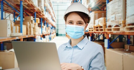 Portrait of female administrator working in warehouse typing text on laptop with helmet and protective medical mask.