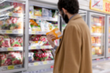A masked man in a supermarket selects an item in the freezer section. Blurred. Coronavirus pandemic.