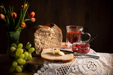 Frühstück ist fertig! Brot ist geschnitten, mit Butter geschmiert, Glas Tee , Weintrauben, Tulpen und Erdbeere Marmelade  stehen daneben auf dem Holztisch  Mit weißen Tischdecke dekoriert. Rustic