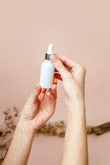 Transparent dropper bottle with serum lotion or essential oil in female hand over beige trendy neautral color background. Skin care cosmetics concept, eart tones, beauty flyer