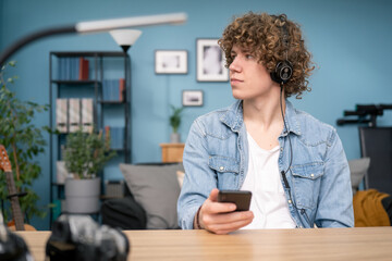 Portrait of Teenager student learning music sitting at desk in his living room at home. The boy wearing blue shirt has headphones on head and holding a smartphone looking window.