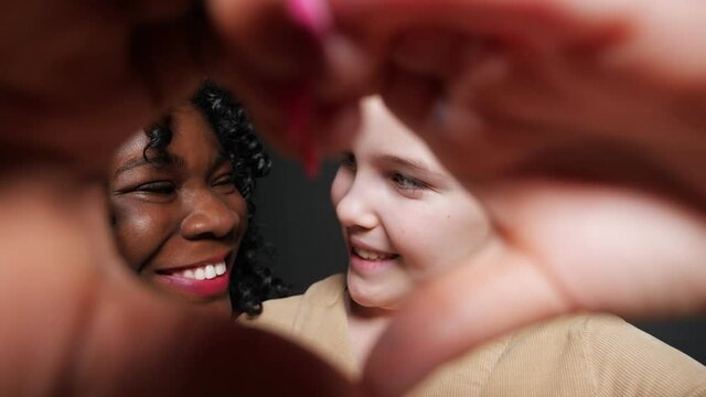 Black stepmother with curly hair and blonde adopted schoolgirl show heart sign with hands and look into eyes with joyful smiles closeup