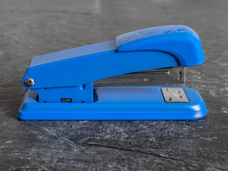 A blue metal office stapler stands on a black concrete background