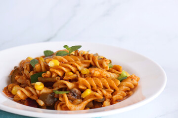 Whole wheat fusilli pasta with vegetables. Vegetable Pasta with zucchini, mushrooms, corn, and peppers on a white plate. Garnished with mint leaves. on a white marble table with cutlery. copy space.
