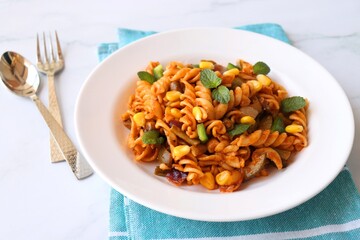 Whole wheat fusilli pasta with vegetables. Vegetable Pasta with zucchini, mushrooms, corn, and peppers on a white plate. Garnished with mint leaves. on a white marble table with cutlery. copy space.