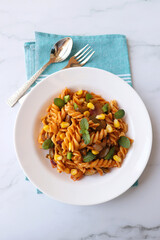 Whole wheat fusilli pasta with vegetables. Vegetable Pasta with zucchini, mushrooms, corn, and peppers on a white plate. Garnished with mint leaves. on a white marble table with cutlery. copy space.