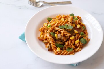 Whole wheat fusilli pasta with vegetables. Vegetable Pasta with zucchini, mushrooms, corn, and peppers on a white plate. Garnished with mint leaves. on a white marble table with cutlery. copy space.