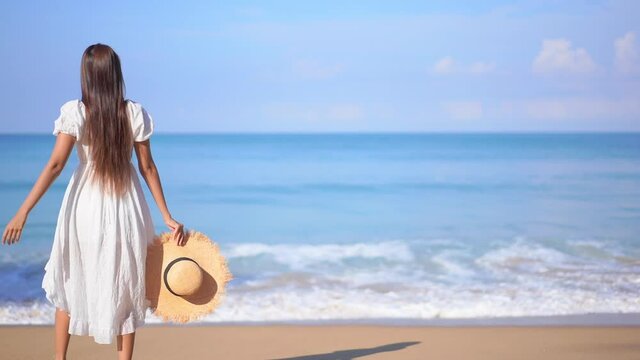 Happy Asian woman in white sundress walking on the white sand beach at seafront raising her hands up turning around enjoying freedom at a tropical island