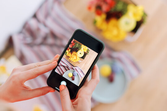 Woman taking photo of flowers and macaroons with smartphone. Instagram photography blogging workshop concept. A girl hanging a phone taking a photo of present on wooden table.