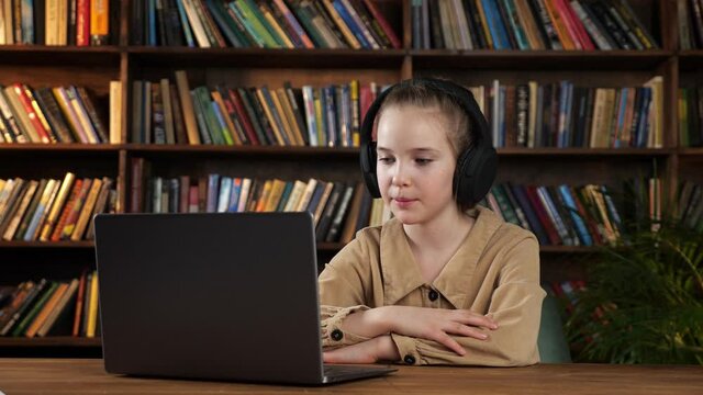 Lady junior student in black headphones listens to teacher and answers sitting at wooden table with grey laptop against racks of books