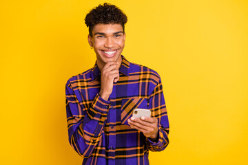 Photo of young afro guy happy positive smile hand touch chin think browse cellphone isolated over yellow color background