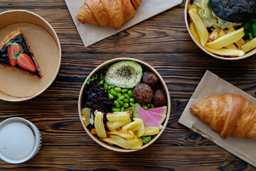 Different takeout food on wooden kitchen table. Hawaiian vegan poke bowl with variety of veggies, french croissant, salmon burger and blueberry cheesecake. Close up, top view, copy space, background.