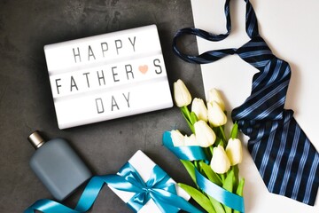 Happy fathers day. Light box with the text HAPPY FATHER'S DAY near a tie, cologne, gift box and a bouquet of tulips on a gray and white background. Holiday concept.
