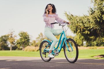 Full size photo of impressed brunette lady ride bicycle talk telephone wear lilac top pants sneakers outside in park