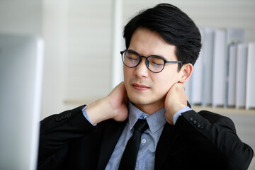 Asian young black short hair male employee worker wears formal suit with necktie gray shirt and eyeglasses has office syndrome hurt shoulder and neck when working in front computer monitor at table