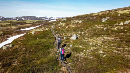 Fototapeta premium A couple hiking through the highlands of Eidfjors, Norway. Both of them carry big backpacks. Hike into the wilderness. Snow covers some parts of the slopes. Dry grass around the trail. Rough hike.