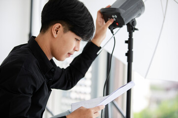 Asian young handsome male short black hair photographer production assistant wears long sleeve shirt and jeans hold document paper in hand while adjusting flashlight in indoor photography studio