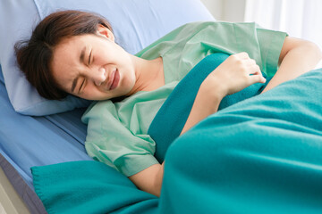 Asian young long hair sick female patient wears green hospital uniform lay down on bed covered by blanket hold hands on stomach twist body hurt and suffering from painful appendicitis in ward room
