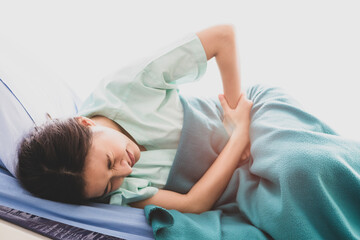 Obraz na płótnie Canvas Asian young long hair sick female patient wears green hospital uniform lay down on bed covered by blanket hold hands on stomach twist body hurt and suffering from painful appendicitis in ward room