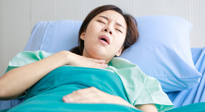 Asian young long hair sick ill girl patient wears green hospital uniform lay down on bed covered by blanket hold hand on chest sudden suffering from cough and pain in private ward room