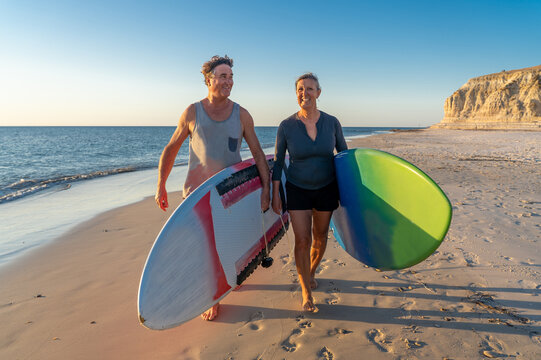 Mature couple surfers with surfboard having fun on empty remote beach enjoying outdoors lifestyle