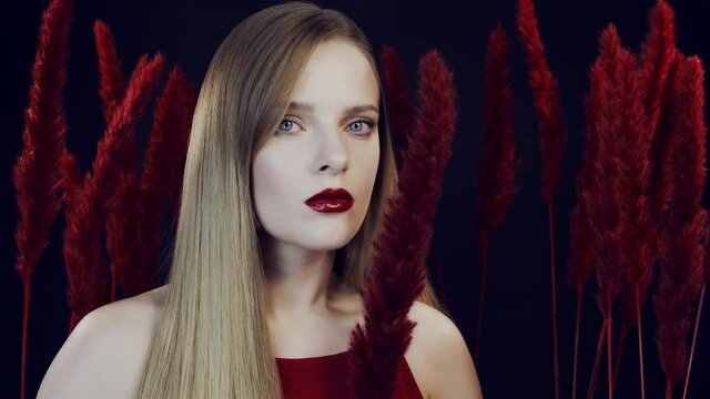 Young woman with red lipstick among red fluffy feathers. The woman looks at the camera. The woman among the racist exotic plants. Blonde with red lips. Woman with a red flower.