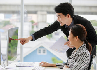 Asian young successful male manager wears black formal suit stand leaning pointing computer screen mentoring and teaching idea to pretty female worker in white stripe casual suit sit learning at desk