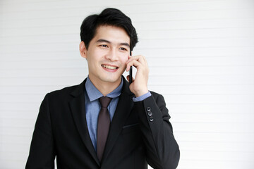 Portrait shot of Asian young successful male ceo businessman wears black formal suit stand smiling talking hold smartphone in hand has conversation with client and customer in front white wall