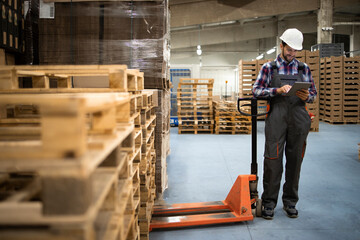 Portrait of warehouse worker typing on tablet computer and standing by wooden palette in factory storage room.