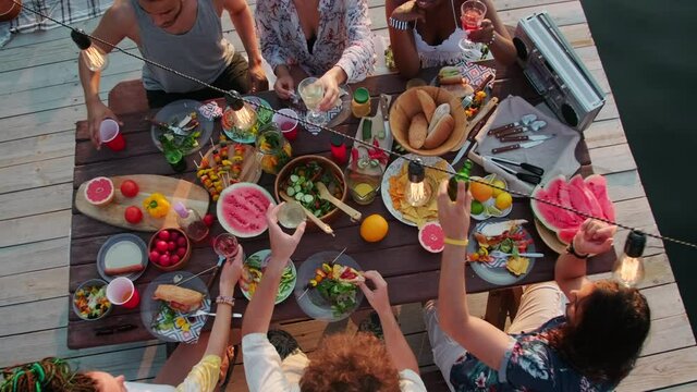 Top down of multiethnic company of young positive friends sitting at dinner table under garland and clinking drinks in toast at lake party on wooden pier