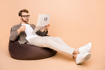 Full size photo of serious focused young man sit chair read paper drink coffee isolated on beige color background