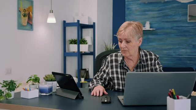 Mature woman using tablet and laptop in same time analysing financial graphs working from home sitting in workplace. Busy focused old employee writing, searching, analysing reports