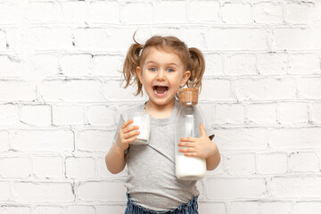 Surprised cute child girl with expression face and mustache of milk on the lips over white brick wall holding glass with milk and bottle of milk.Banner, copy space for text, mock up
