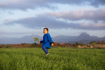 Naklejka na ściany i meble Happy boy walks across a green field with his arms spread and yellow flowers bouqet