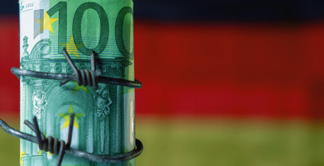 One hundred EURO bancnotes wrapped in barbed wire against flag of Germany as symbol of global economic crisis and recession. Selective focus on money. Copy space for design.