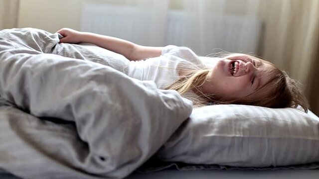 portrait of little blonde girl lying on a grey bed playing hide and seek with blanket and laughing. slow motion