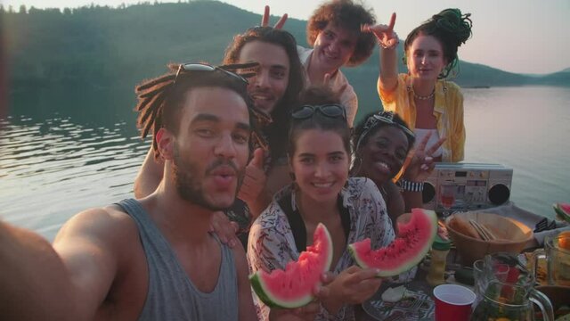 POV slow motion shot of company of young multiethnic friends smiling, waving, eating watermelon and posing for camera while taking selfie together at lake party in summer evening