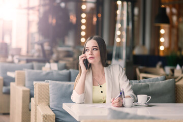 Young woman in white jacket working in cafe with papers, drinking coffee