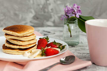 A stack of pancakes with a pink tea mug and  fresh strawberries on a white plate on a light background. A small bouquet of lilacs in a glass vase stands next to the Breakfast.