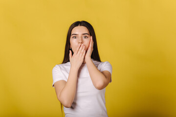 Surprised girl close mous by hands. Attractive young female in studio on yellow background. Image of excited young woman standing isolated