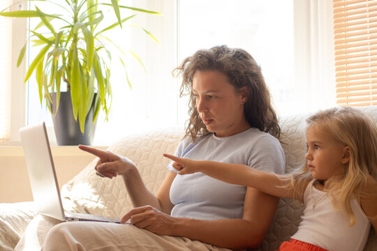 Single mother teaching little daughter using laptop computer, pointing at screen sitting on sofa at home.Babysitter showing training materials, watching cartoon with pupil, online educational program