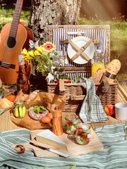 Vintage picnic basket, hamper with baguette and lemonade outdoors on a grass with cheese, mozzarella, tomatoes, cherries, vine. Guitar, snacks on straw mat. Eco friendly picnic outdoors.