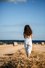 Fototapeta na wymiar Portrait of a young curly-haired woman in a wheat field, where wheat is mowed and sheaves are standing, enjoying nature. Nature. sun rays Agriculture