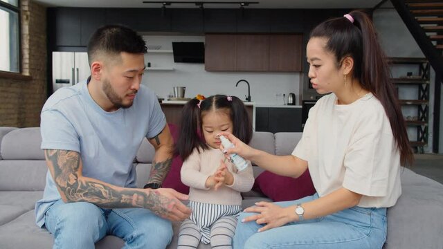 Positive caring handsome asian parents teaching adorable preschool age daughter to hygiene, disinfecting hands with hand sanitizer while spending leisure together , relaxing on sofa at home.