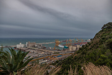 View of the port of Barcelona before the storm. Dramatic sky, on the horizon the sea with waves. In the foreground are green trees and bushes.