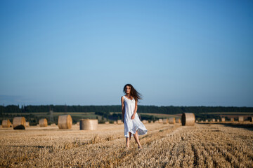 Naklejka na ściany i meble A young beautiful woman in a white summer dress stands on a mown wheat field with huge sheaves of hay, enjoying nature. Nature in the village. Selective focus